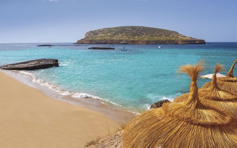 Playa de arena clara y aguas turquesas en Ibiza, con sombrillas de fibras naturales en primer plano y una pequeña isla frente al horizonte. La escena transmite verano, descanso y belleza mediterránea, ideal para representar un día de playa o una experiencia junto al mar.