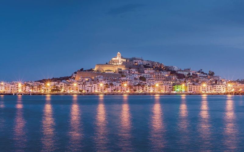 Vista panorámica de Dalt Vila y el puerto de Ibiza al anochecer, con las luces de la ciudad reflejándose sobre el mar en calma. La imagen transmite una Ibiza histórica, elegante y mediterránea, perfecta para representar la isla desde una perspectiva tranquila y luminosa.