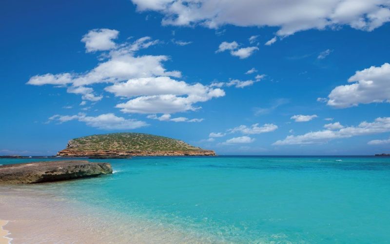 Playa de aguas turquesas y arena clara en Ibiza, con cielo azul, nubes suaves y un paisaje mediterráneo ideal para disfrutar del mar.