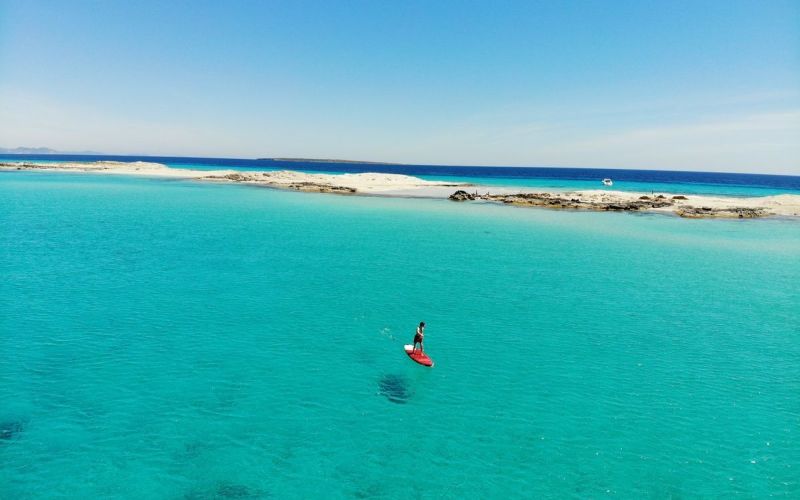 Aguas turquesas de Formentera con una persona practicando paddle surf, arena blanca y horizonte abierto en un paisaje mediterráneo tranquilo.
